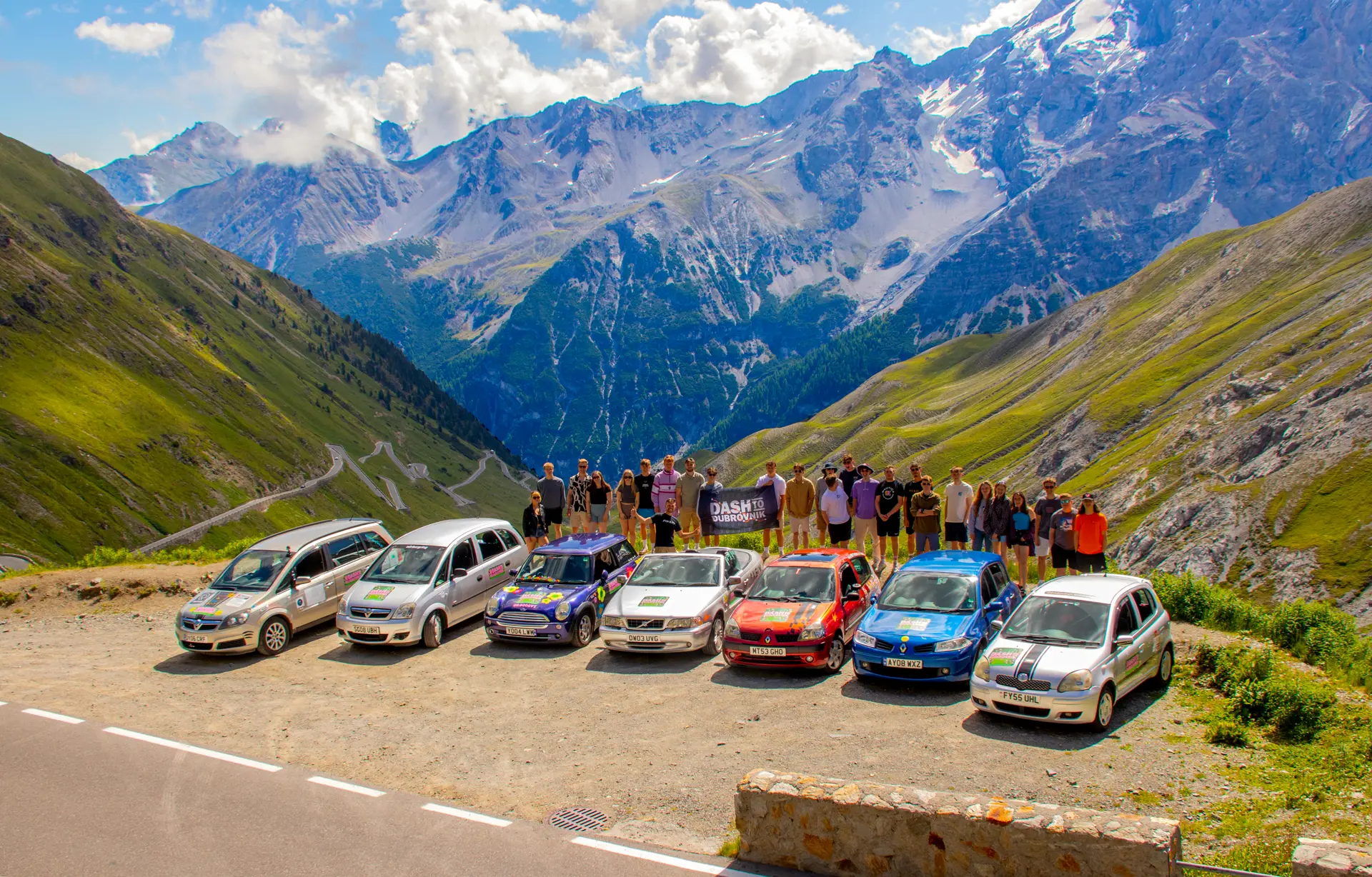 Dash to Dubrovnik convoy of decorated banger cars and the full team holding the Dash banner at an alpine mountain pass.