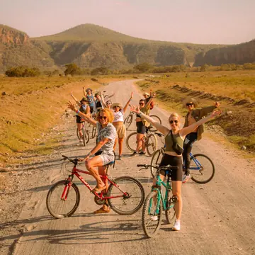 Group on bikes waving on a dirt road with hills behind on the SYB Kenya trip