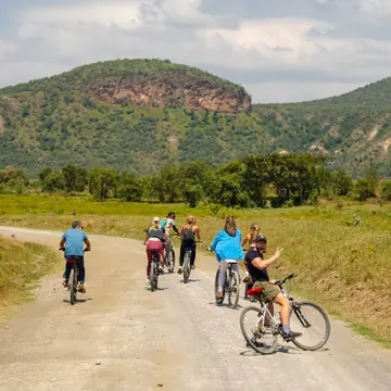 Cyclists riding a dirt road toward a green flat topped hill on the SYB Kenya trip