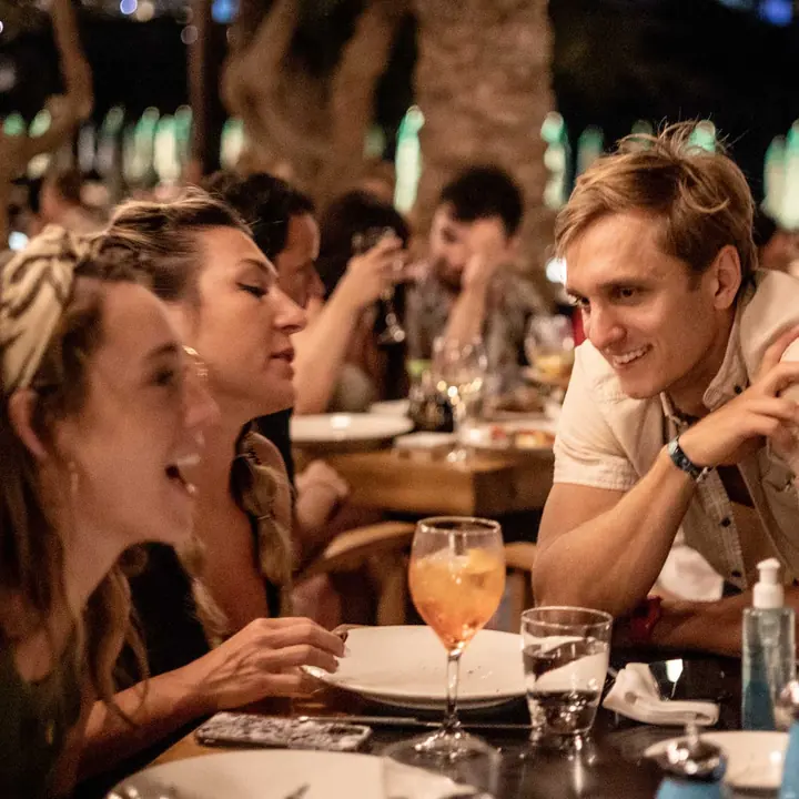 Friends laughing over dinner and drinks at a restaurant table at night on the SYB Greece trip