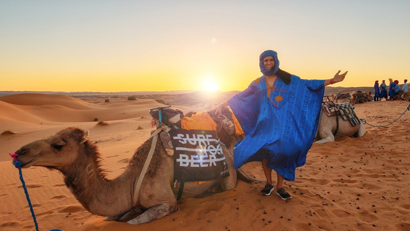 Mantas in a blue robe next to a camel in the Moroccan desert at sunset