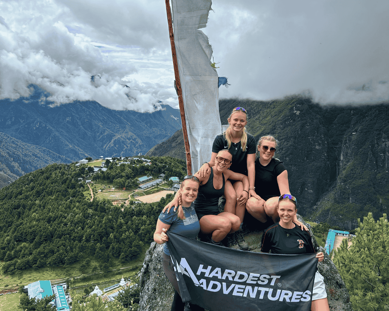 Group of female hikers sitting on a rock holding a Hardest Adventures banner with a view of the Himalayas in the background on their way to everest base camp.