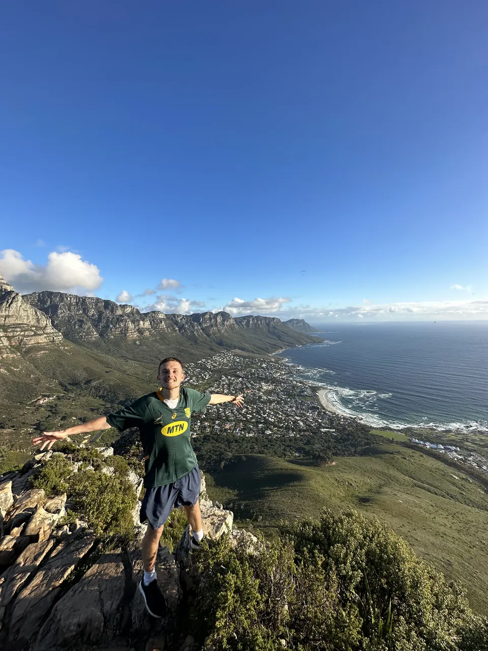 Bailey Turvey with his arms out on a rocky ridge, the sea and a coastal town far below.