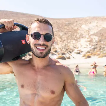Carrying the speaker on his shoulder beside clear turquoise water on the SYB Greece trip