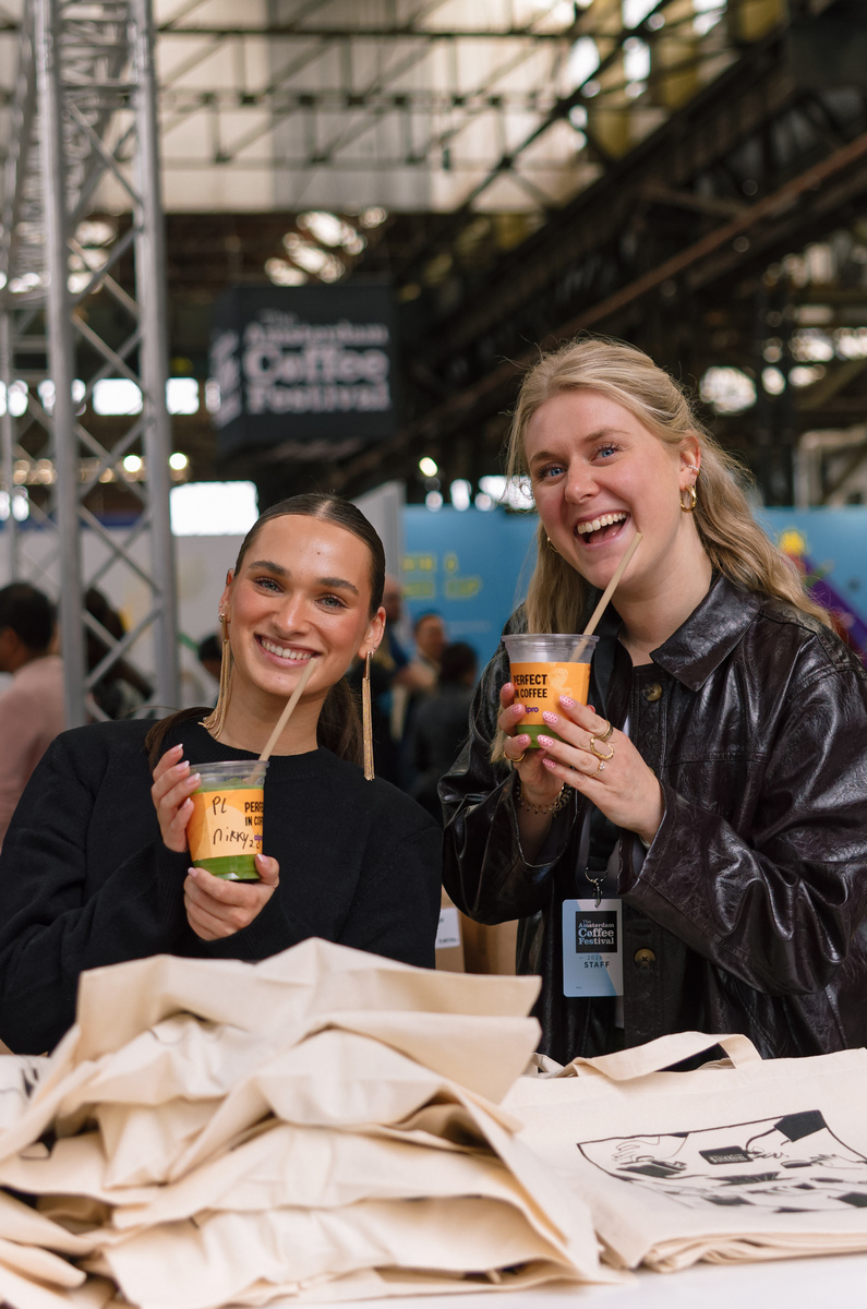 Two smiling ACF crew members holding iced coffees behind a stack of festival tote bags