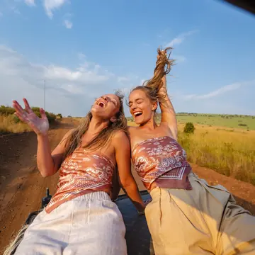 Two friends laughing on the roof of a safari jeep under a blue sky in Kenya