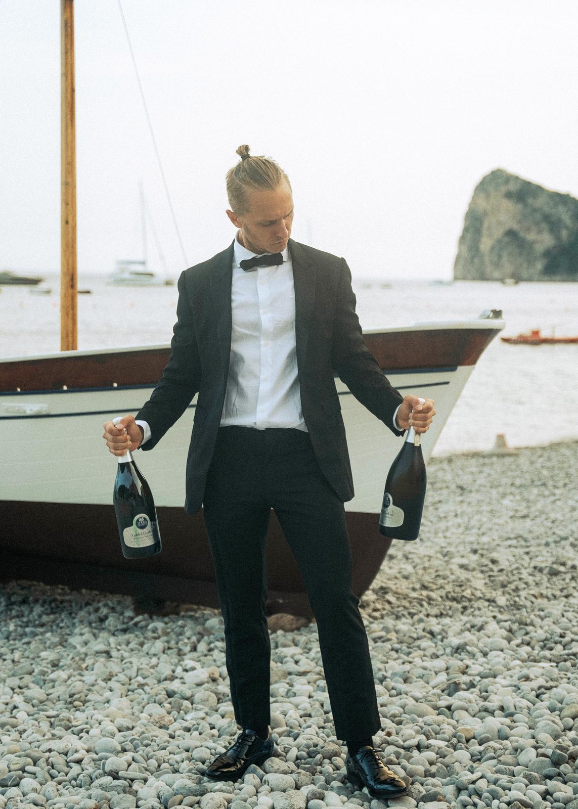 Mantas in a tuxedo holding two bottles of bubbly on a beach on the Amalfi Coast