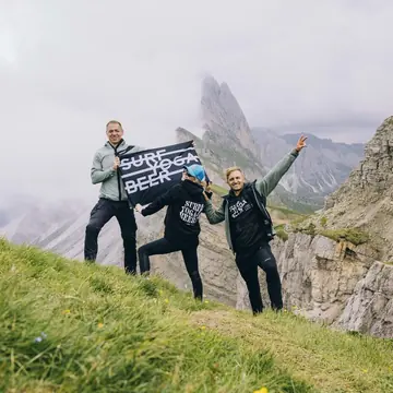 Three hikers holding the Surf Yoga Beer flag in front of jagged peaks on the SYB Dolomites trip
