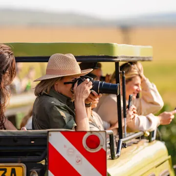 Guest in a sun hat photographing wildlife from the open roof of a safari jeep in Kenya