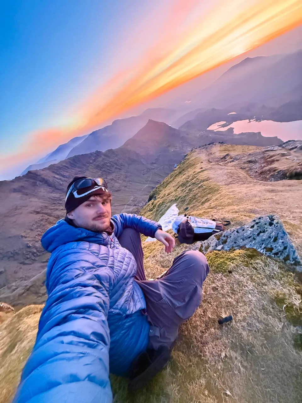 Tom Savage taking a selfie on a rocky summit at sunset, peaks and cloud glowing pink behind him.