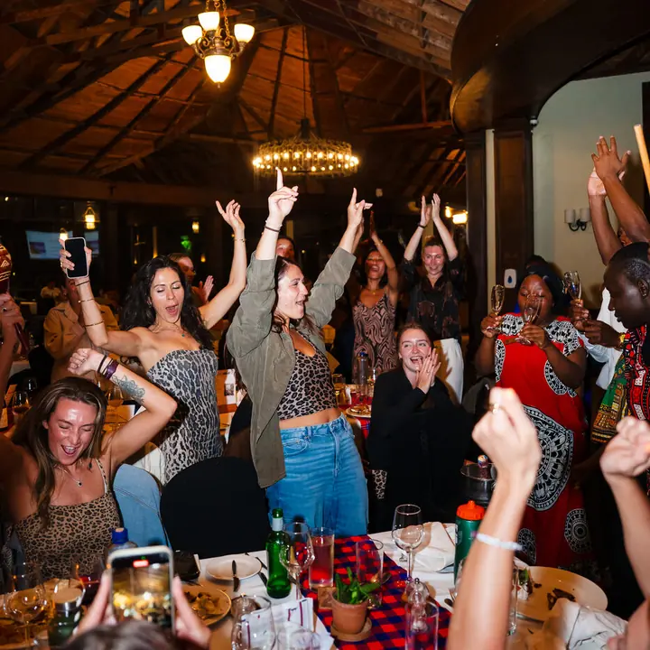 The crew dancing with arms in the air around the dinner table at the lodge in Kenya