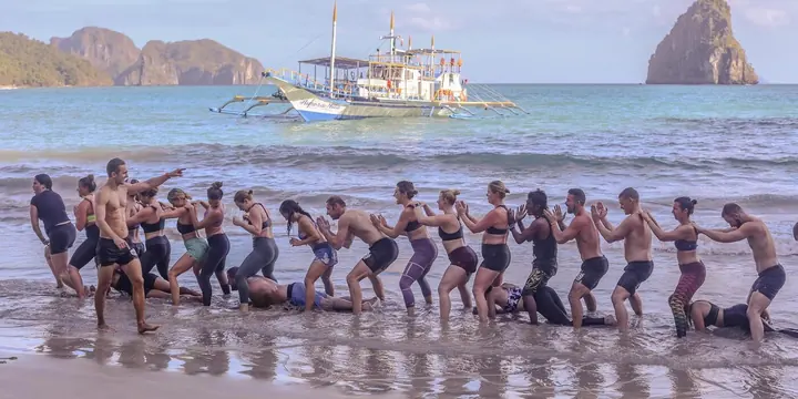 The whole group in a line in the shallows with an outrigger boat behind on the SYB Philippines trip