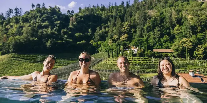Four friends in the outdoor pool with vineyards and forest behind on the SYB Dolomites trip