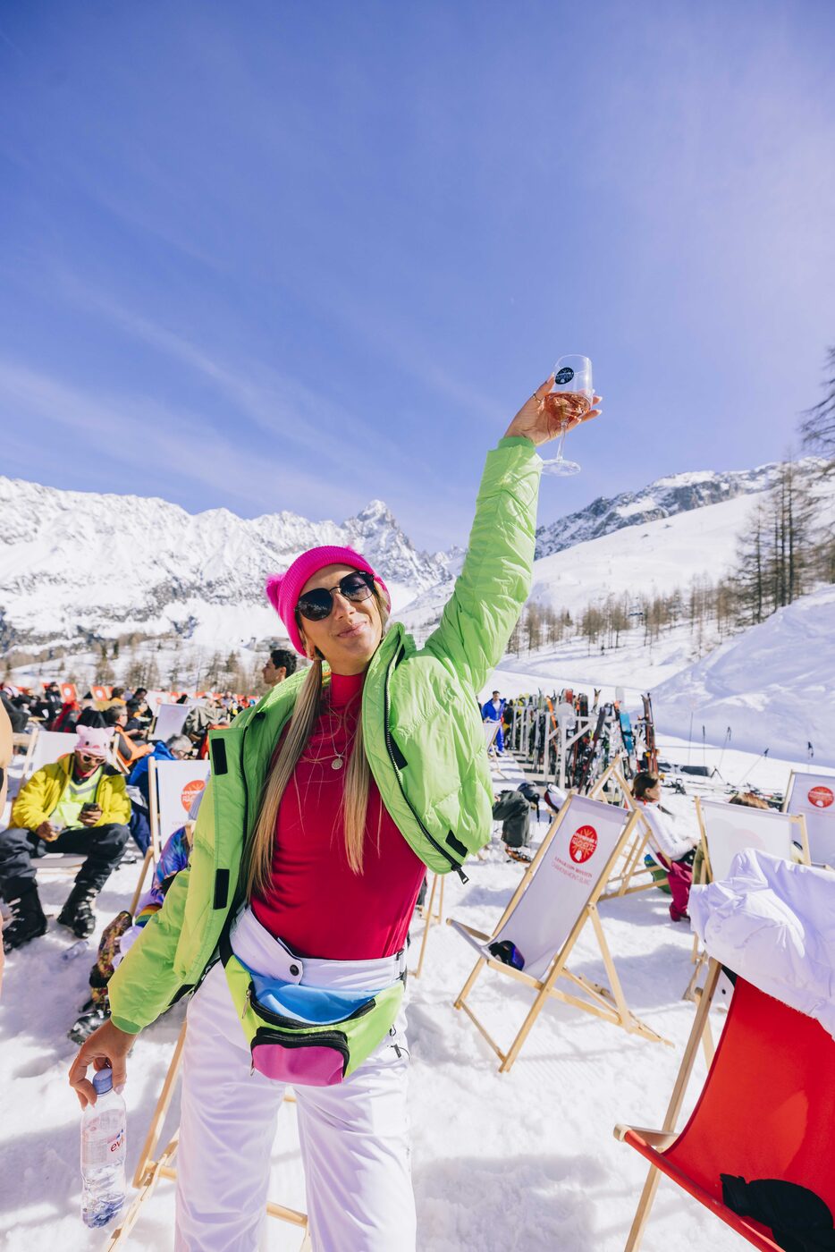 A skier in a green jacket cheering on a sunny mountain terrace
