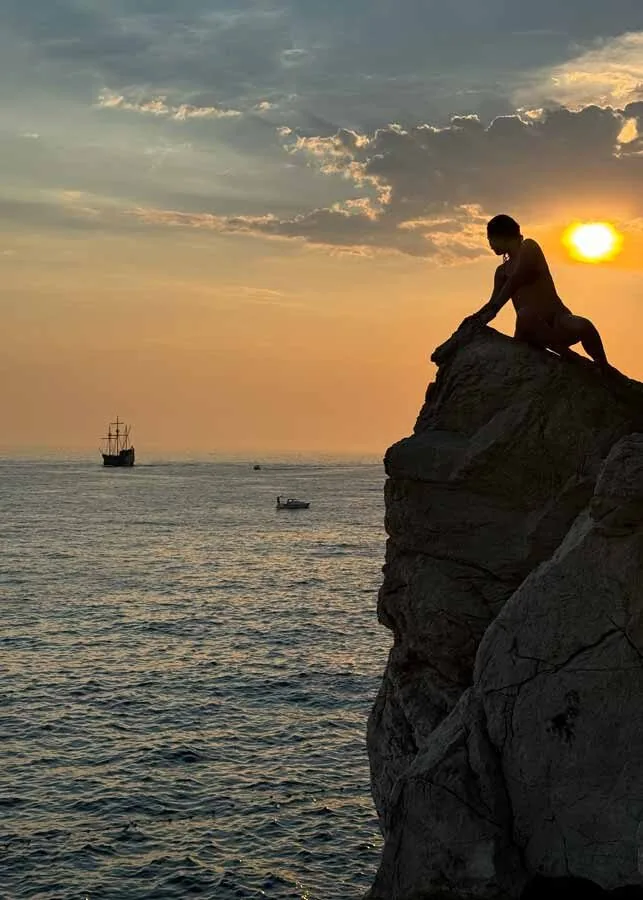 A traveler sitting on a cliff edge at sunset with a ship out on the water in Croatia