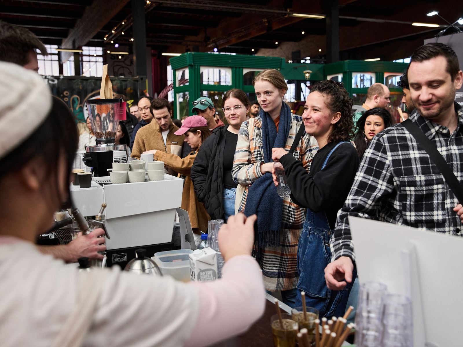 A barista hands out coffee to a smiling crowd at a roaster's bar inside the NDSM hall
