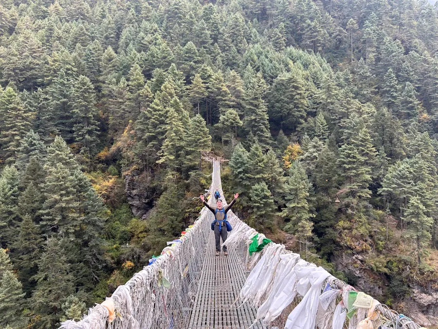 Trekker crossing a high suspension bridge with arms out wide on the Everest Base Camp trek with Hardest Adventures.