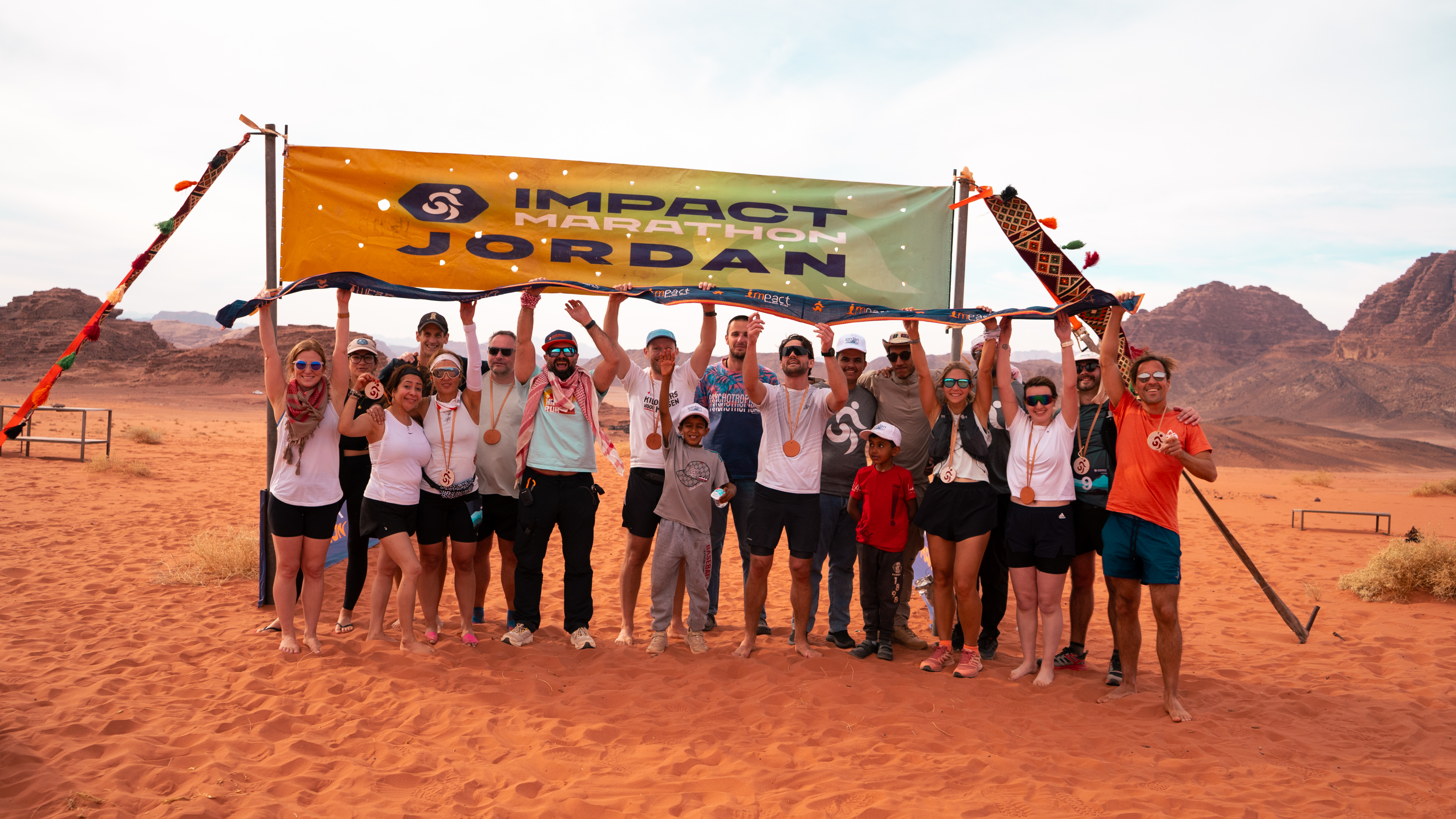 A diverse group of runners and local community members stand together celebrating at the Impact Marathon Jordan finish line in the desert. They are holding up a large orange "Impact Marathon Jordan" banner against a backdrop of rugged mountains and sand.