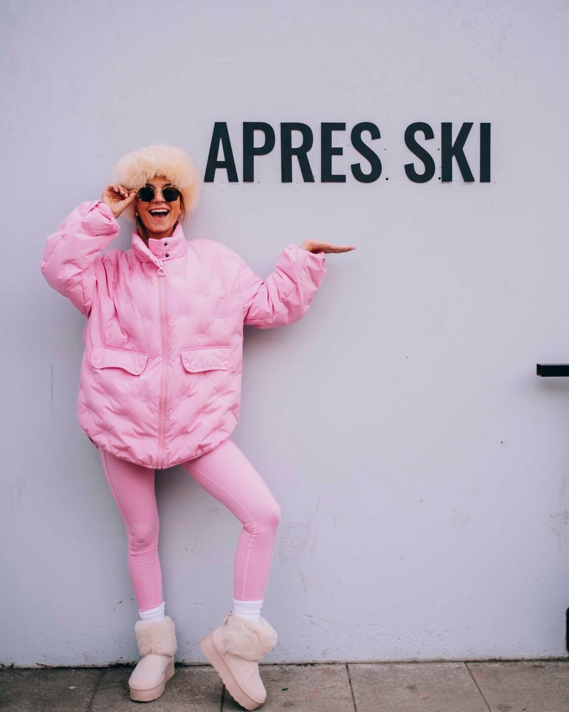 A traveler in a pink ski outfit posing next to a sign that says apres ski
