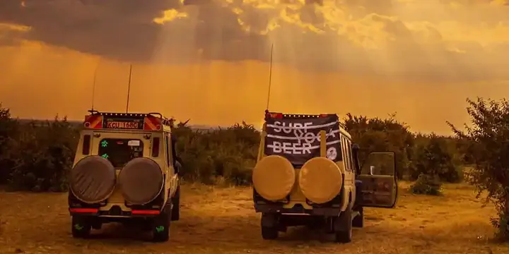 Two safari jeeps parked under golden sunset rays, one flying the Surf Yoga Beer flag, in Kenya