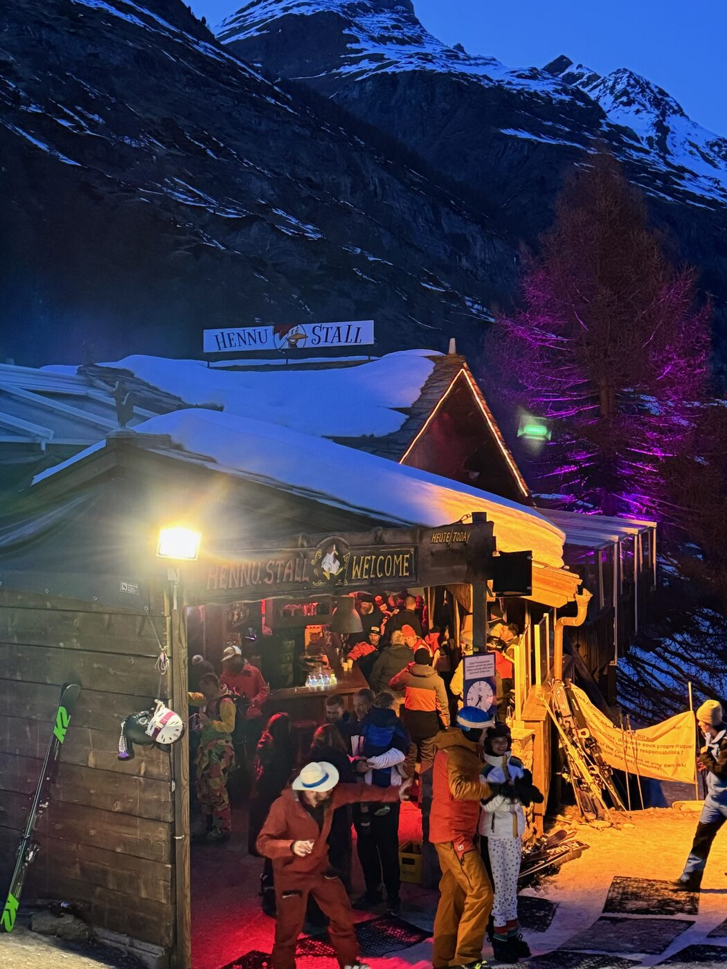 A crowd at an aprรจs ski bar lit up at night in the snow