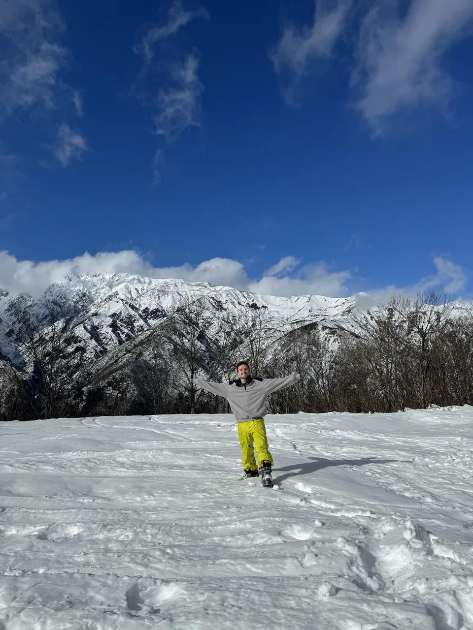 Bailey Turvey with his arms out on a snowfield, snow-capped mountains and a blue sky behind him.