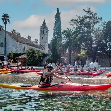 Sea kayaker paddling past a row of kayaks and a stone church on the SYB Croatia trip