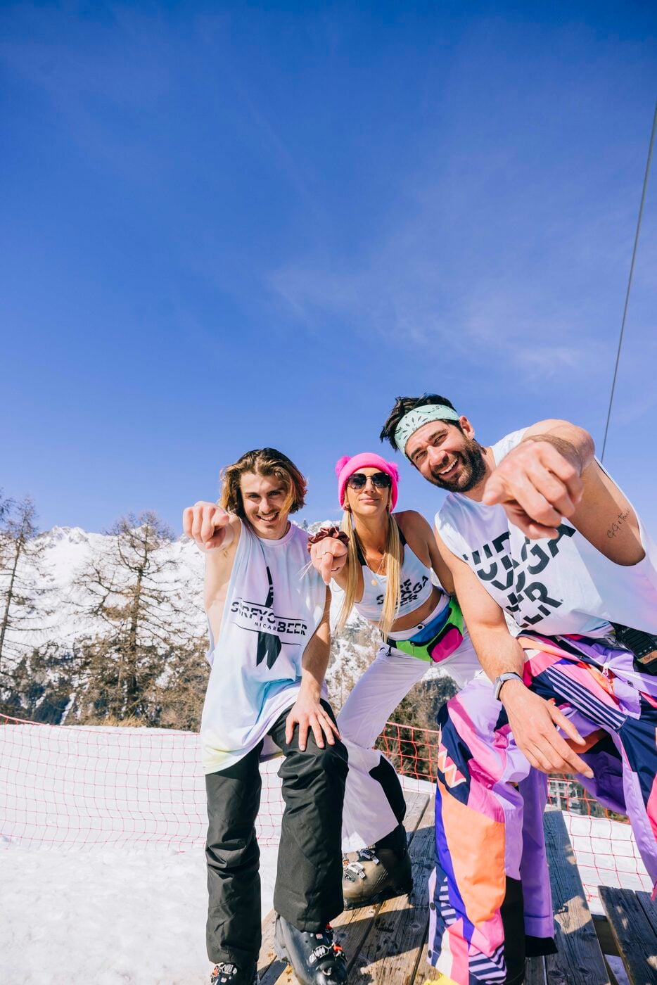 Three friends in SYB shirts laughing on the snow under a blue sky