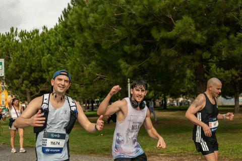 Two runners laughing and joking for the camera mid-race on a tree-lined marathon route.