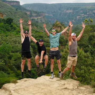 Four friends jumping on a rock ledge above a green gorge on the SYB Kenya trip