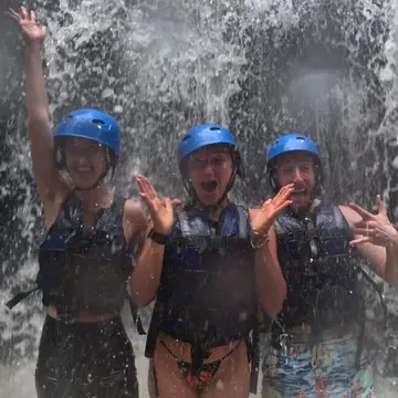 Three friends in helmets standing under a crashing waterfall on the SYB Bali trip