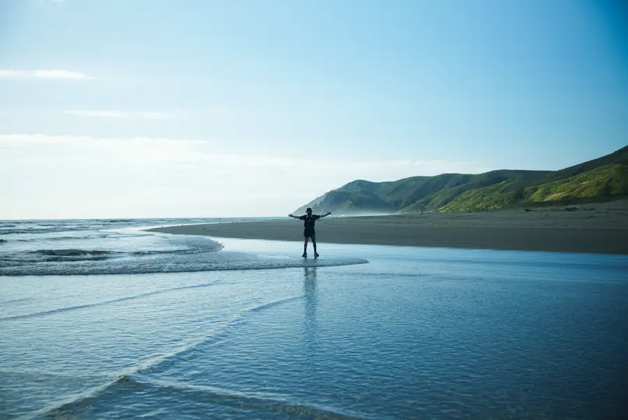 A lone figure standing with arms outstretched on vast tidal flats at low tide, beneath a coastal headland in New Zealand