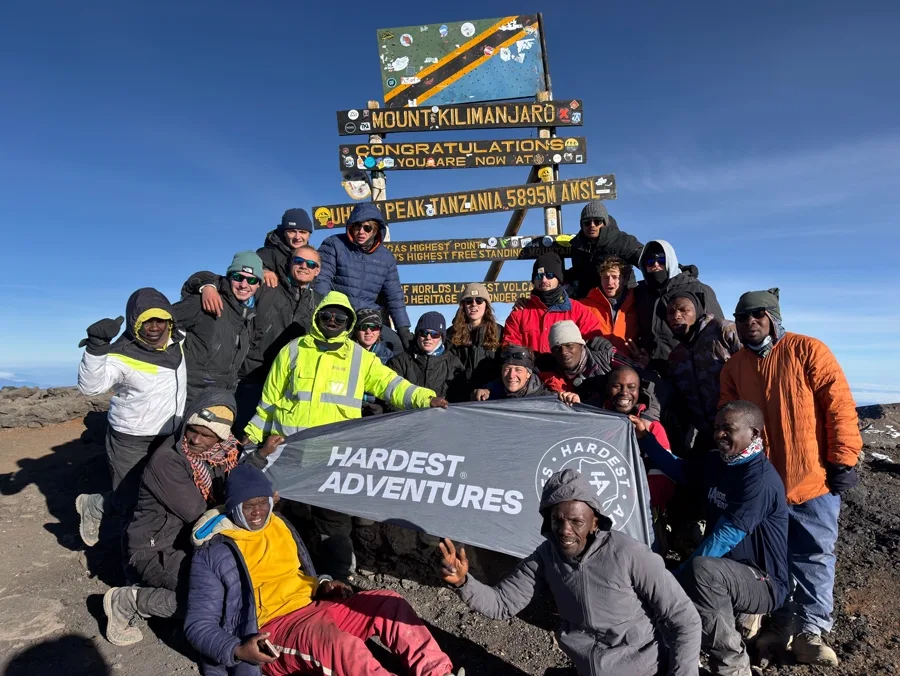 Hardest Adventures group and their Kilimanjaro guides holding the HA banner at the Uhuru Peak summit sign