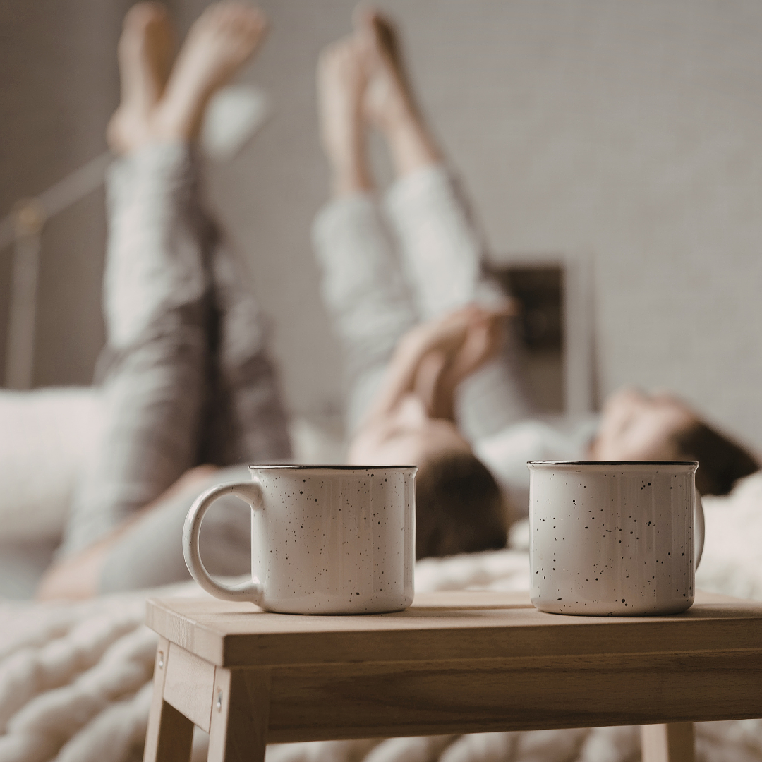 Twoceramiccoffeemugsonawoodenbedsidetablewithacouplerelaxinginbedinthebackground