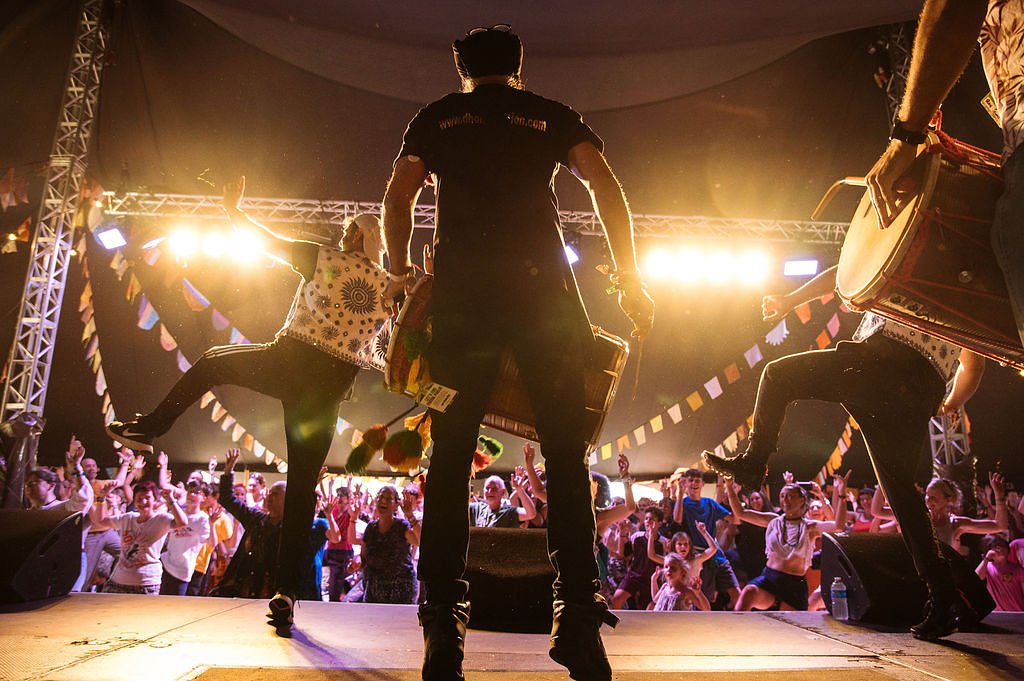 3 people on an elevated stage. One person plays a Dhol, while the other two demonstrate Bhangra dance moves to the audience, who copy their movements. 