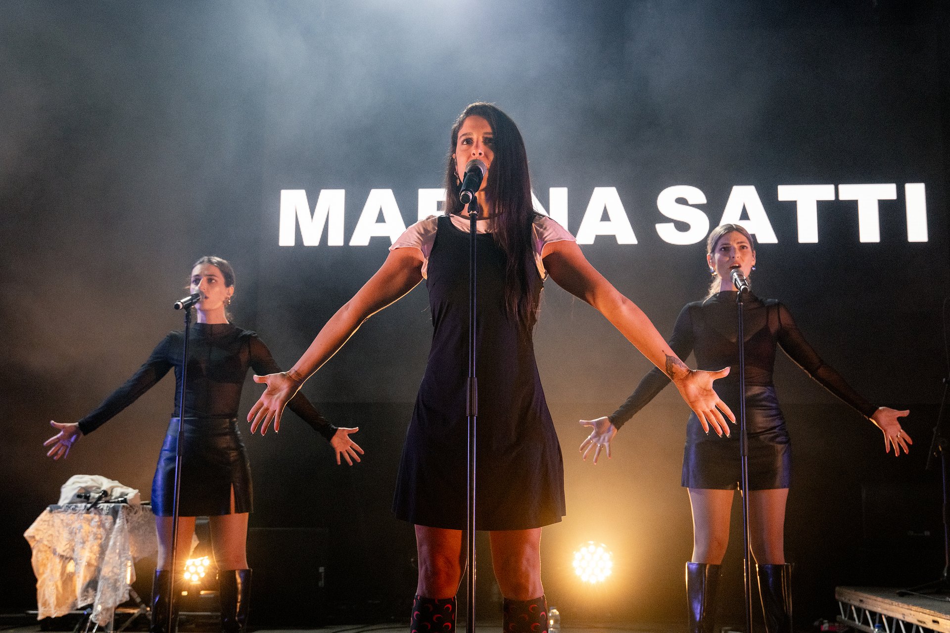 Marina Satti performing, she stands centre stage wearing a black dress with her arms out by her side. Her backing singers are stood behind mimicking her position. 