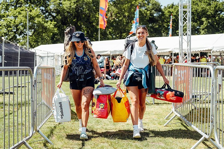 Two girls on sports tour walk through the entrance carrying their camping equipment at Bournemouth 7s Festival