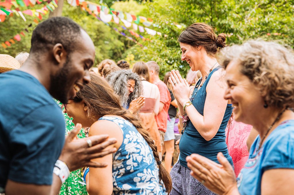 A close up image of two people looking at each other and bowing in a yoga class. They hold their hands together in front of them in a prayer position, and are smiling at each other as other people do the same in the background. 