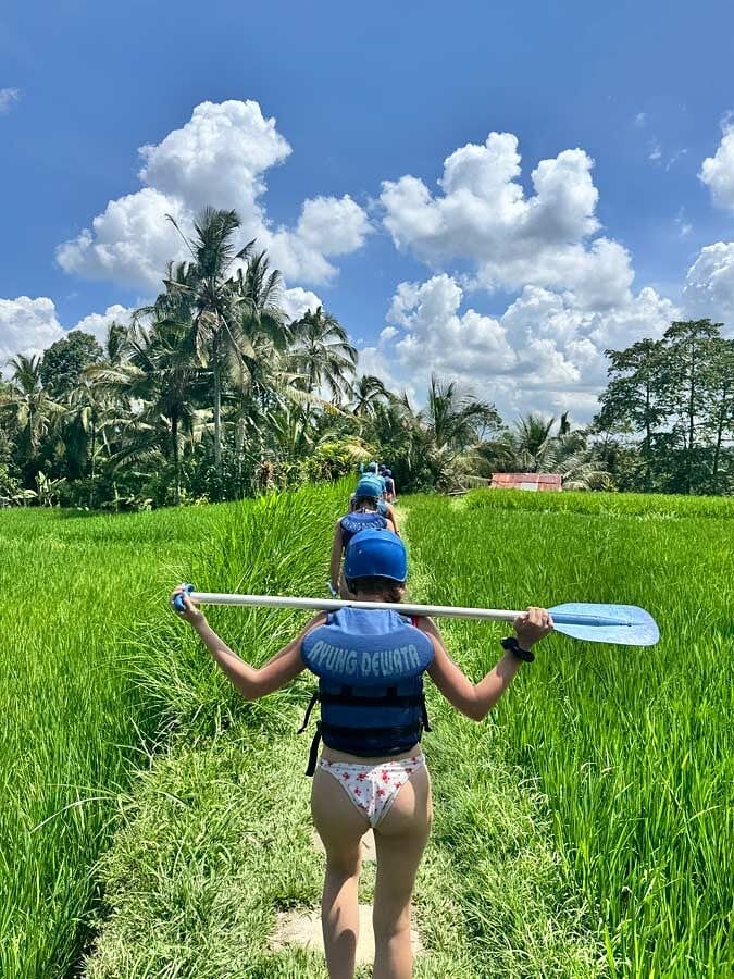 A traveler in a life vest carrying a paddle through green rice fields in Bali