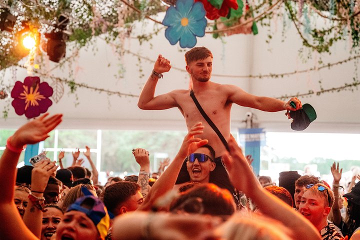 Man on friends shoulders dancing in a large music crowd in the tropical VIP area at Bournemouth 7s Festival, during a tribute act, in Dorset England.