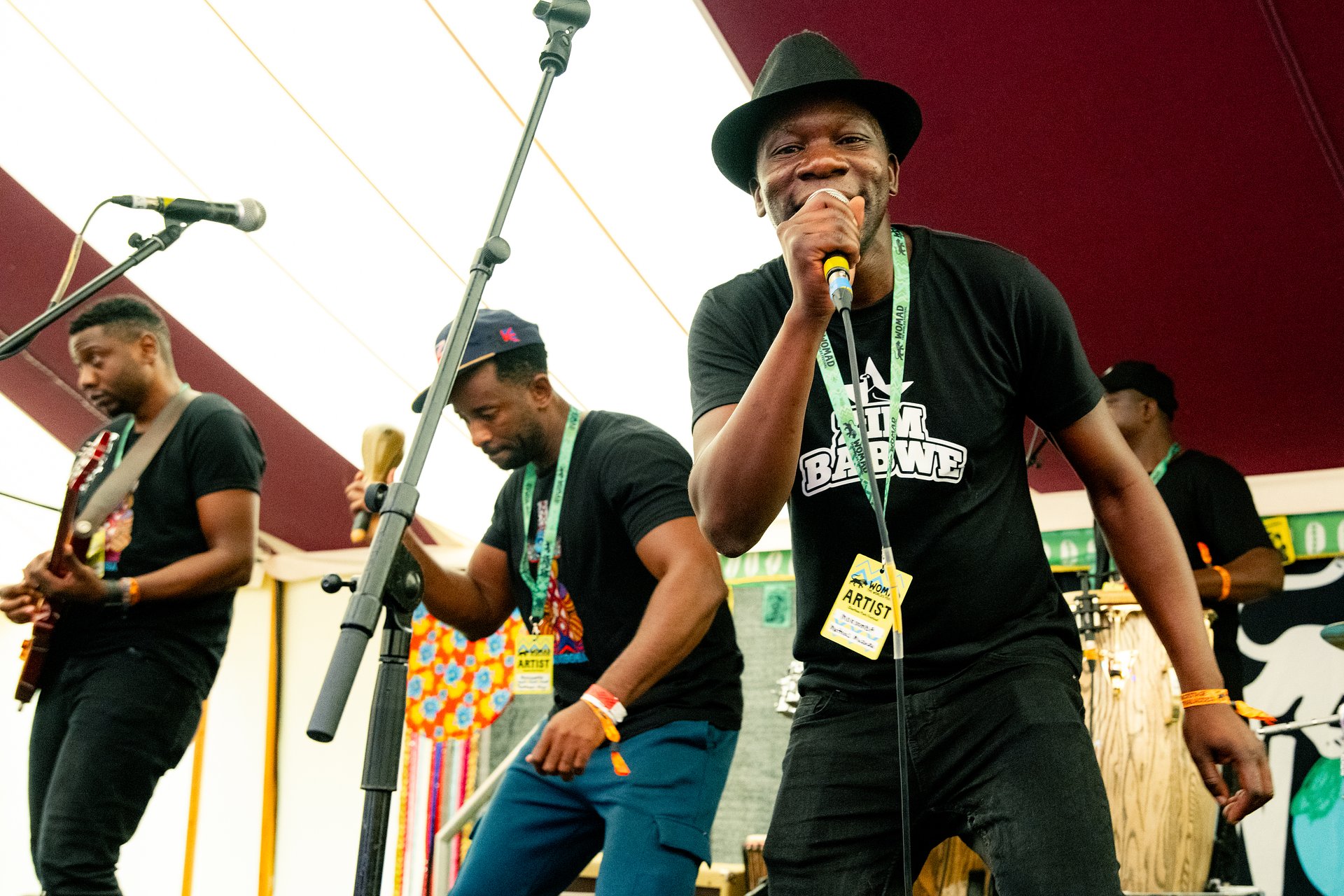 Band members of Mokoomba performing in the Workshops tent. The main singer looks into the camera holding a microphone, and the drummer, percussionist and guitarist all play their instruments. 