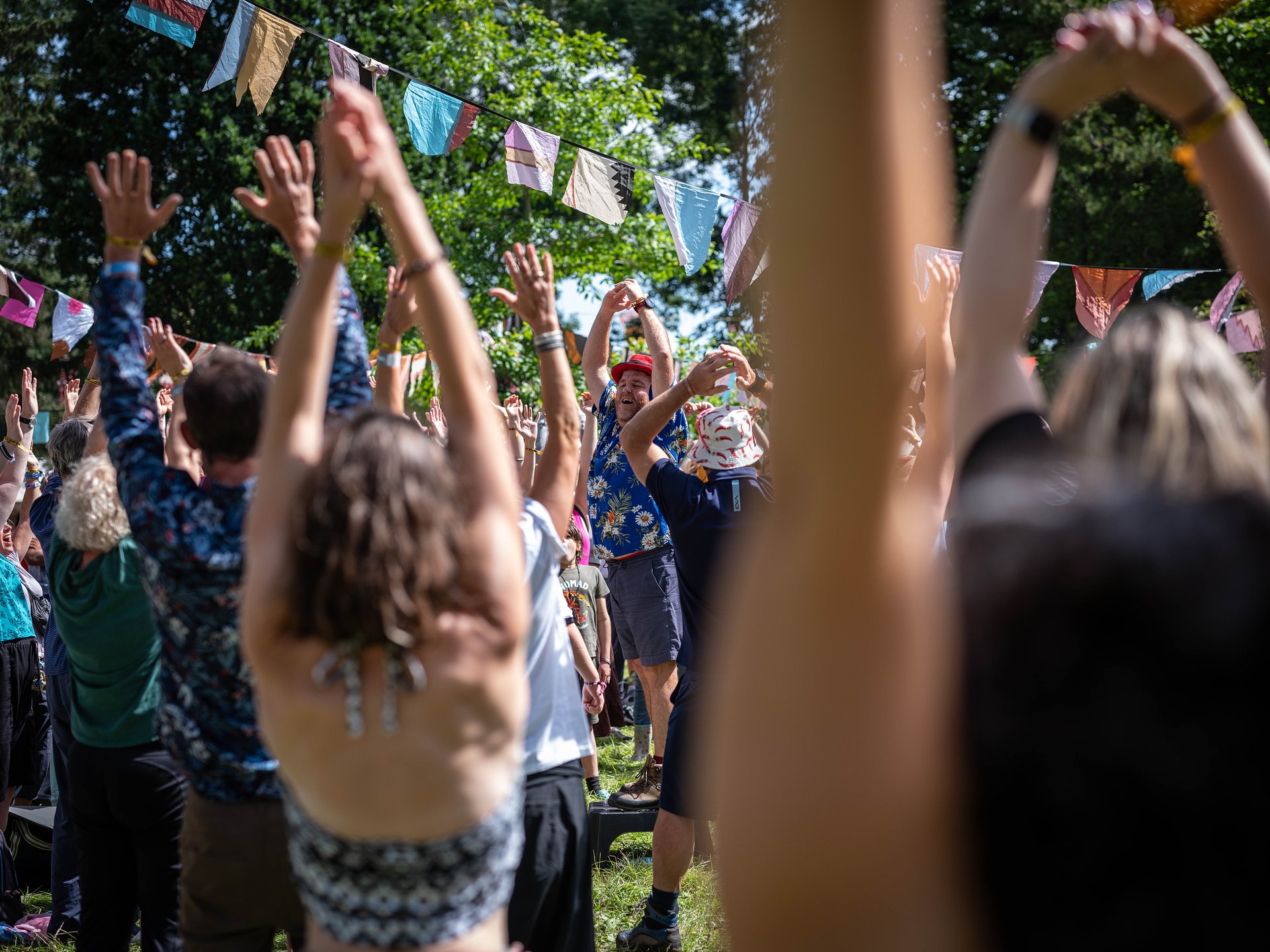 People taking part in a yoga class in the sun. Participants have their arms lifted above their heads, copying the yoga instructor in the middle of the image. 