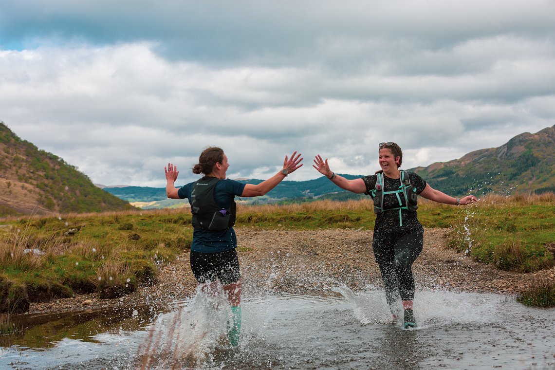Two women, Sophie Power from SheRACES and Anna from The Running Channel, celebrating together as they splash through a stream during the Scotland Impact Marathon, symbolising camaraderie and empowerment in trail running.